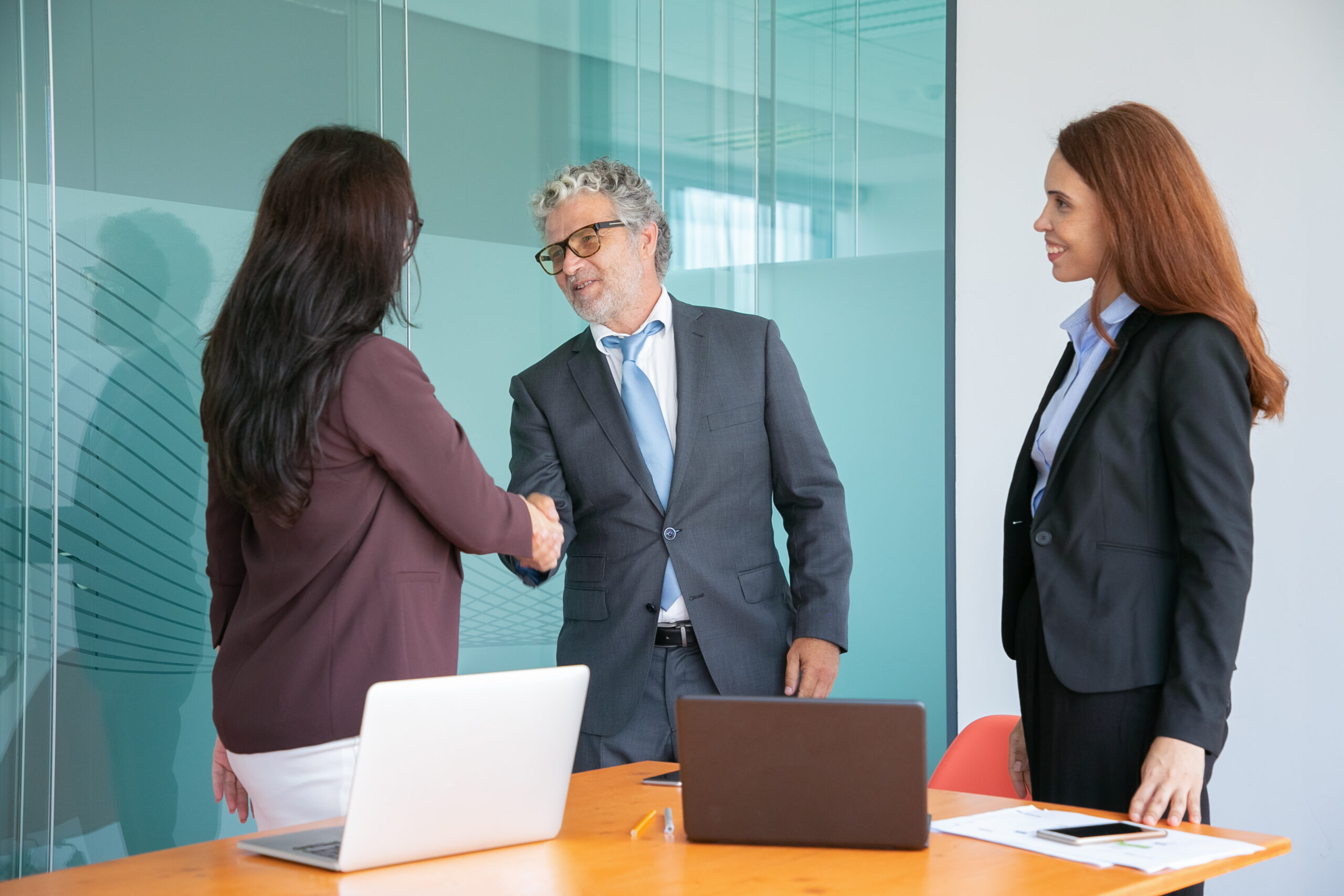 Grey-haired senior manager handshaking and greeting businesswoman. Red-haired professional businesswoman standing, smiling and looking at partners. Business, collaboration and meeting concept